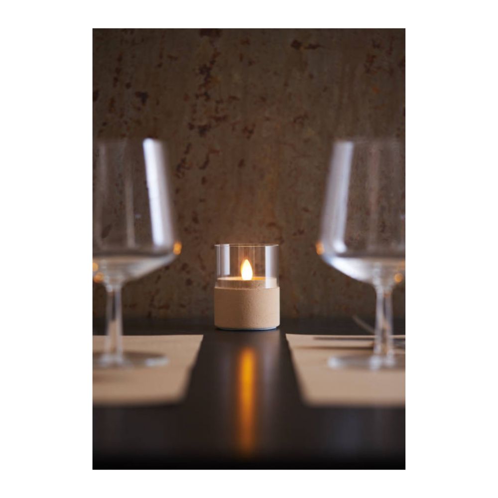 A LED Moving Flame (38x36 mm) candle from Duni GmbH flickers realistically on a dark table between two empty wine glasses and beige placemats in front of a brown, textured wall.