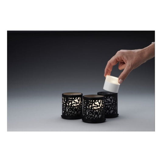 Eine Hand legt eine LED aus dem Duni LED-Kerzen-Set in eine schwarze Fassung. Daneben leuchten zwei ähnliche Fassungen mit aufwendigen Ausschnitten. Der Hintergrund weist einen Farbverlauf von Dunkel- zu Hellgrau auf.