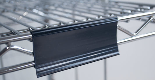 Nahaufnahme eines Novocal Etikettenrahmens mit Papierschildern (ca. 100 x 44 mm, 1 Stück), der an einem Metalldrahtregal befestigt ist, mit dem schwarzen Kunststoffetikettenhalter an der Vorderkante; der Hintergrund ist unscharf.
