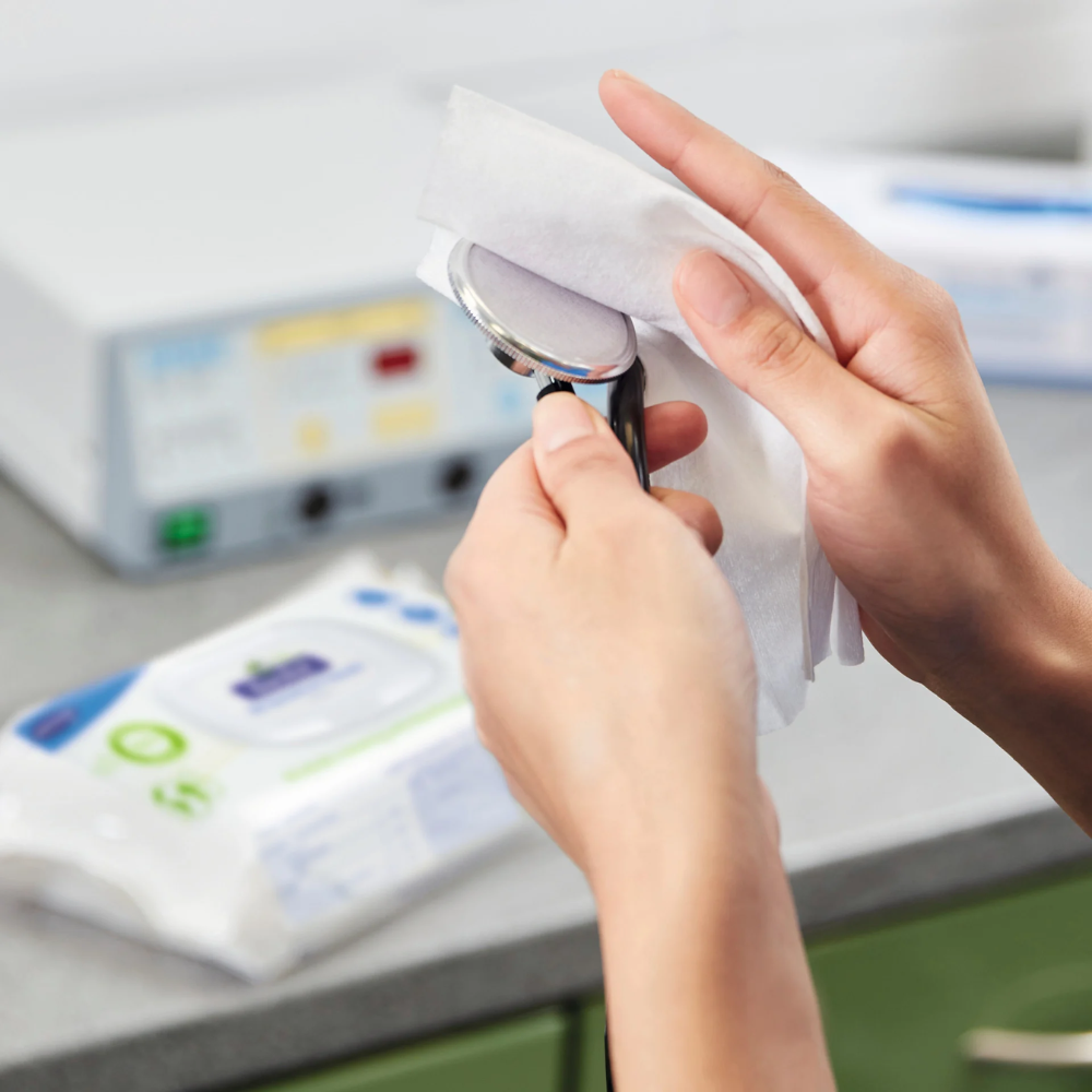 A person cleans a medical instrument with Hartmann Bacillol® 30 Sensitive Green Tissues Rapid Disinfection Wipes from Paul Hartmann AG. In the background, a pack of disinfection wipes and various medical devices can be seen. The setting appears to be a clean, clinical environment, emphasizing the use of environmentally friendly, material-friendly disinfection wipes.