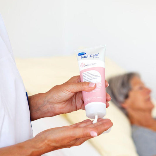 Eine Person gibt Hartmann MoliCare Skin Hautschutzcreme (200 ml) der Paul Hartmann AG, angereichert mit Nutriskin Protection Complex, in ihre Handfläche. Im unscharfen Hintergrund liegt eine kleine, grauhaarige Person auf einem Bett und vermittelt so den Eindruck von persönlicher Pflege oder einem medizinischen Umfeld.