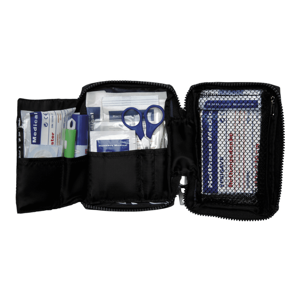 A Holthaus Medical first aid kit TRAVEL blue/red with zipper from Holthaus Medical GmbH & Co. KG is open and shows various first aid items. The items include scissors, plasters, alcohol swabs, gauze compresses, and a green crayon. A mesh pocket on the right side contains additional plasters and a first aid brochure for quick reference.