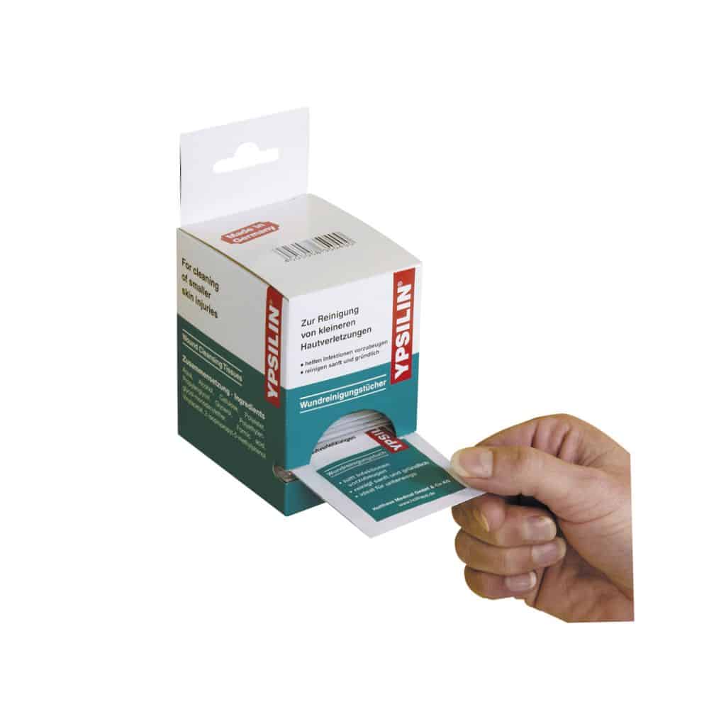 Eine Hand zieht ein Holthaus Medical YPSILIN® Wundreinigungstuch aus einem Kartonspender. Die Verpackung trägt einen deutschen Text und die Schachtel ist weiß und grün, mit deutlich sichtbaren Produktinformationen und dem Markennamen von Holthaus Medical GmbH & Co. KG.