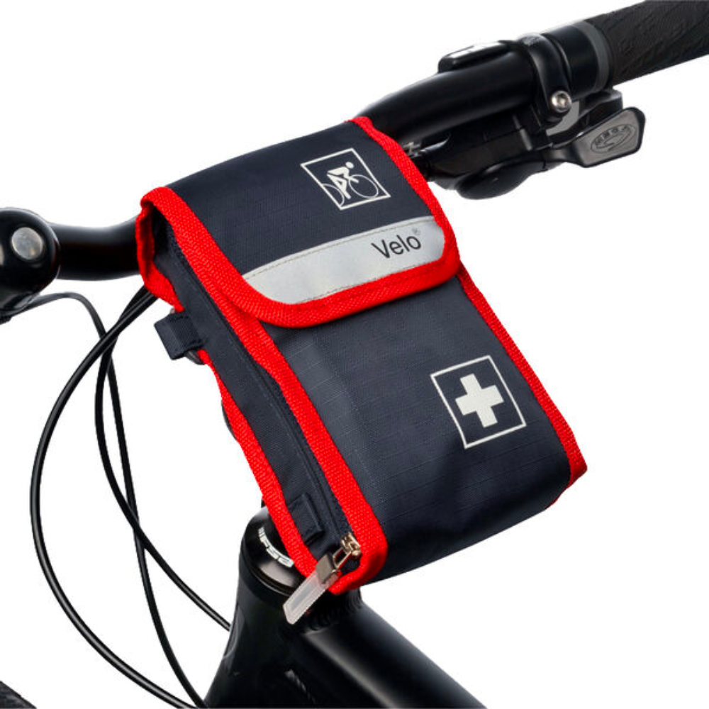 Ein kleines, schwarz-rotes Erste-Hilfe-Set mit der Aufschrift „Holthaus Velo® Fahrradverbandtasche - 21-teilig | Packung (1 Stück)“ der Holthaus Medical GmbH & Co. KG ist an der Lenkstange eines Fahrrads befestigt. Die Verbandtasche des Fahrrads ist mit einem weißen Kreuzsymbol und einem Fahrradsymbol versehen. Zu sehen sind außerdem die Lenkstange und ein Teil des Fahrradrahmens.