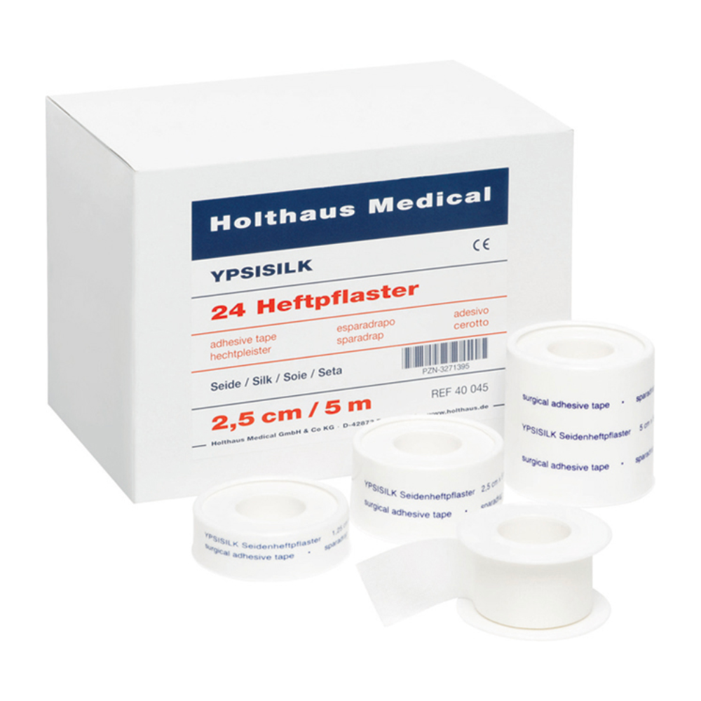 A box labeled „Holthaus Medical GmbH & Co. KG“ with „Holthaus YPSISILK Adhesive Plaster – 2.5 cm x 5 m | Package (5 m)“ and several rolls of white tape in different sizes in front. The text on the box and the tapes contains product details in multiple languages.