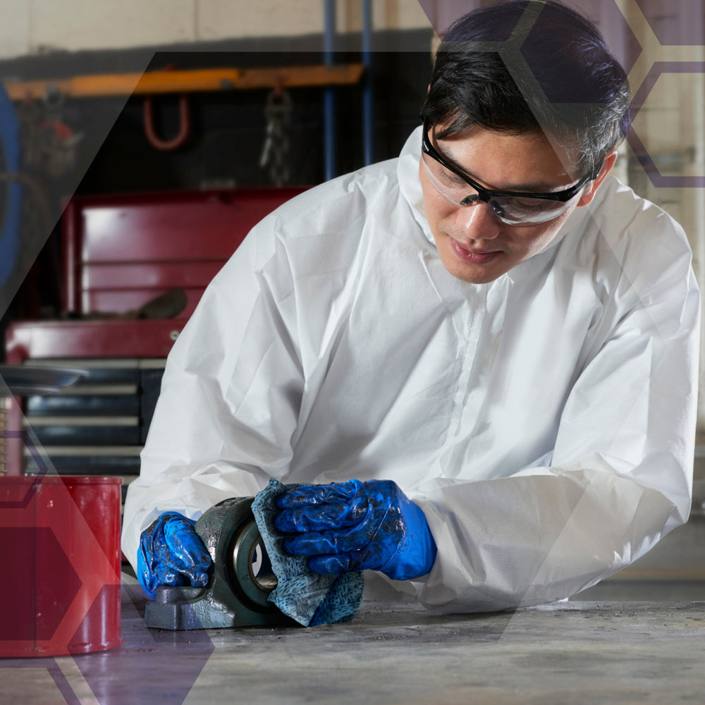 In einem weißen Anzug, mit blauen Handschuhen und einer Schutzbrille kniet eine Person auf dem Boden und verwendet Kimtech® Prozeßwischtücher - Großrolle (blau) von Kimberly-Clark GmbH, um ein mechanisches Teil in einer industriellen Reinigung zu reinigen.