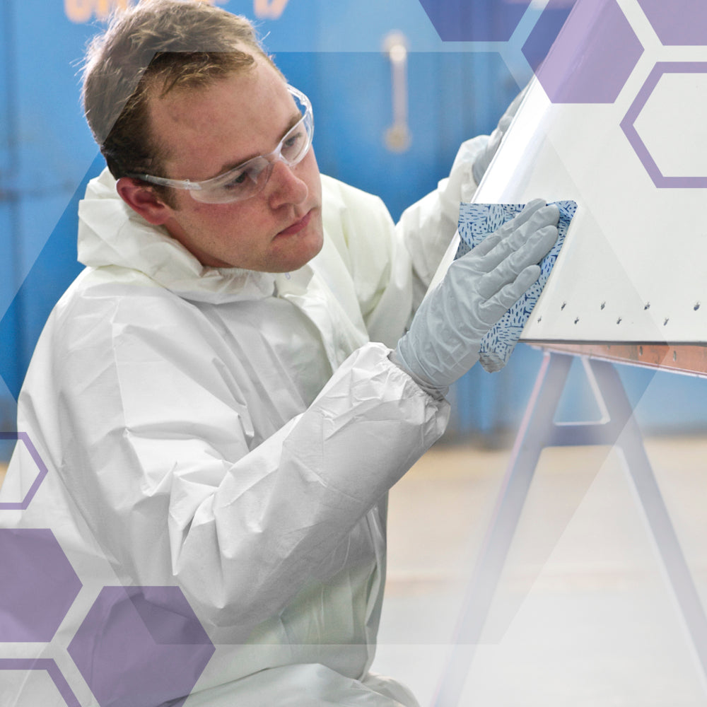 Eine Person mit Schutzbrille, Handschuhen und weißem Anzug verwendet Kimberly-Clark GmbH Kimtech® Prozeßwischtücher (BRAG™ Box, Blau, 160 Tücher), um ein weißes Objekt in einem Innenraum zu reinigen, wobei lila Sechsecke auf eine innovative industrielle Reinigung hinweisen.