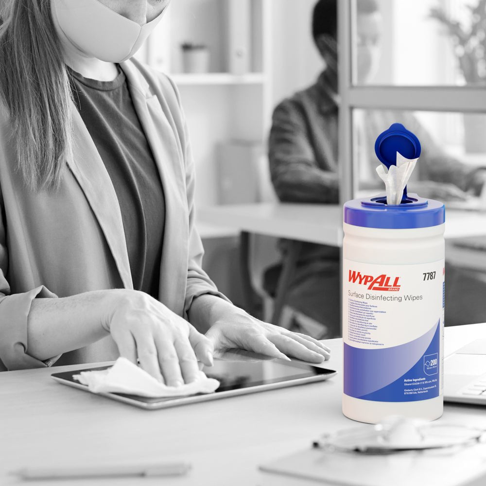 A masked person disinfects a tablet at a desk with WypAll® surface disinfection wipes (dispenser bucket, 200 sheets) from Kimberly-Clark GmbH, with the tray visible, while another person works at a computer in the background.