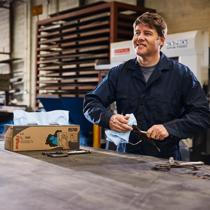 A man in a dark blue overall wipes a tool with a blue disposable towel in an industrial workshop. Next to him on the workbench is a WypAll® X80 PowerClean BRAG™ Box from Kimberly-Clark GmbH (White, 1 Box x 160 Sheets).