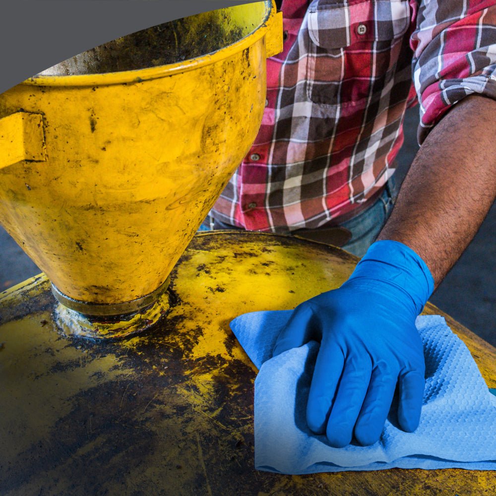 A person in a checkered shirt and blue gloves cleans a yellow industrial machine with the Kimberly-Clark GmbH WypAll® X80 PowerClean - large roll (31.00 cm x 31.50 cm), demonstrating professional use in industry or workshop.