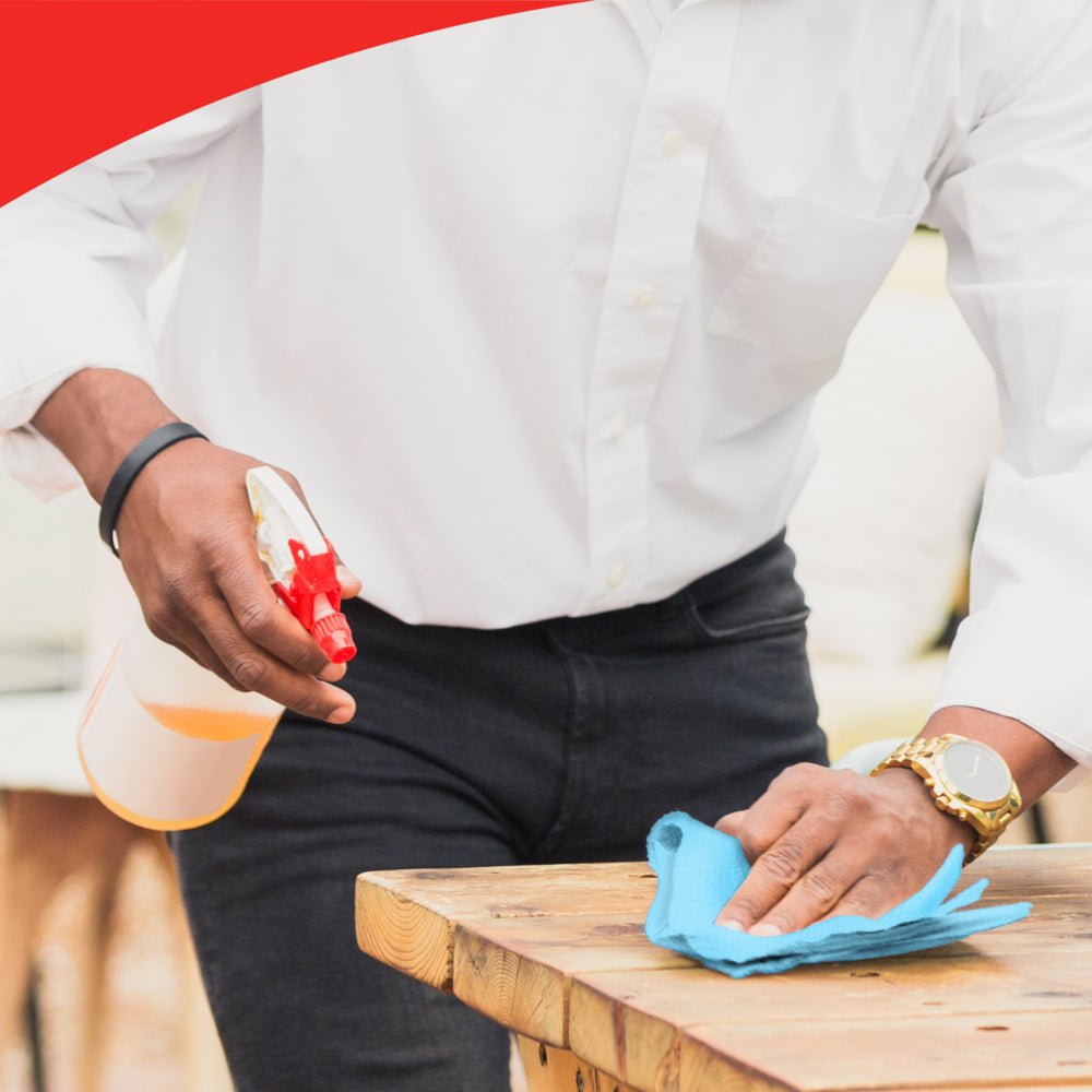 A person with a white shirt and gold watch cleans a wooden table with a blue WypAll® X60 General Clean Roll from Kimberly-Clark GmbH. Their face is not visible. The image has a red border at the top edge.