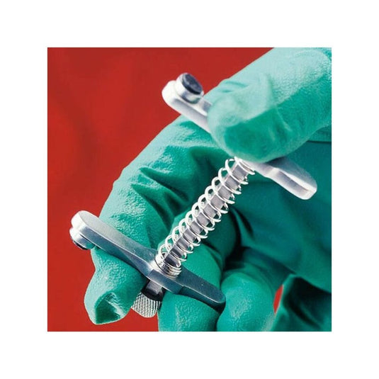 A person wearing green gloves is holding, against a red background, an Altruan Servoprax muzzle for cats with a cylindrical handle and a threaded screw, which is typically used by veterinarians for veterinary procedures.