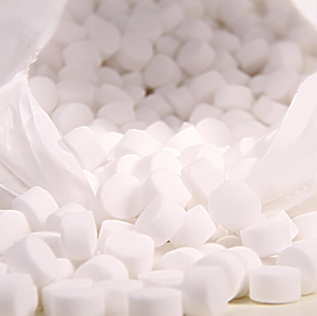 A close-up shows a white plastic bag with the inscription “K+S Minerals and Agriculture GmbH,” containing Regenit salt tablets according to EN 973 Type | Bag (25 kg) that fall onto a surface. The small, round tablets have a uniform shape and size and are ideal for use in water softening systems.