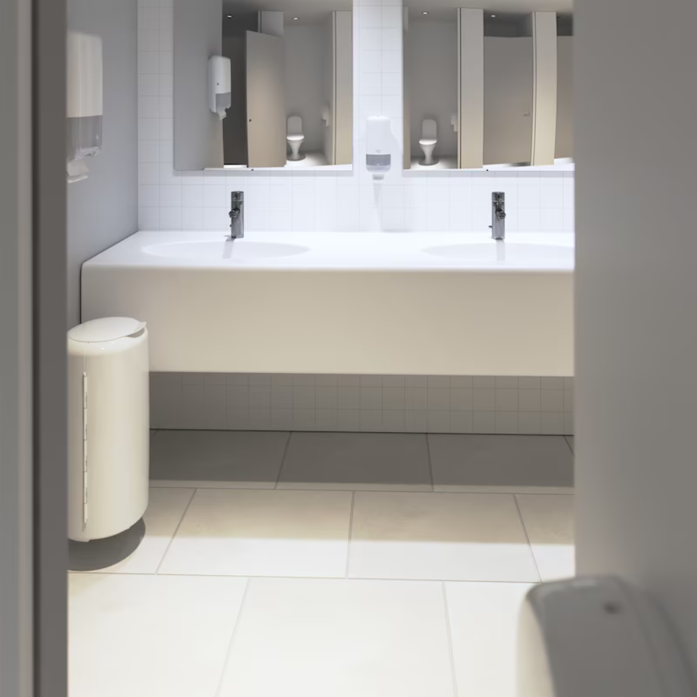 Eine moderne öffentliche Toilette mit einer weiß gefliesten Rückwand und großen Spiegeln über einem Doppelwaschbecken. Über jedem Waschbecken befinden sich Tork 561500 Spender für Seife und Händedesinfektionsmittel Elevation S4 | Packung (1 Stück) Spender von TORK. Im Vordergrund und seitlich sind ein Händetrockner und ein Mülleimer zu sehen, die für optimale Händehygiene sorgen.