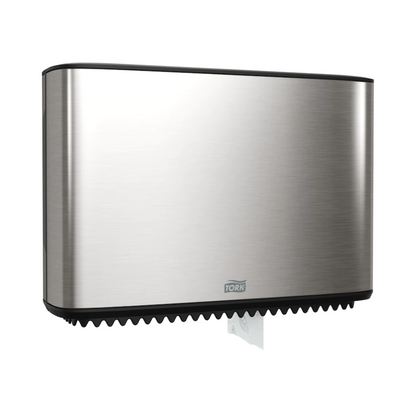 A Tork Xpress dispenser for mini-jumbo toilet paper, part of the Image Line, is elegantly mounted on the wall. This stainless steel dispenser has a sleek rectangular design, with a small section of white paper visible at the bottom. The brand "TORK – Essity Professional Hygiene Germany GmbH" is clearly visible on the front.