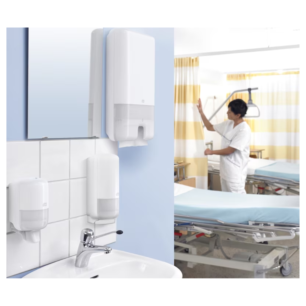 Ein Krankenhauszimmer mit einem Waschbecken, über dem ein Tork Xpress® 471093 Multifold-Handtücher Universal H2 1-lagig | Karton (20 Packungen) und ein Händetrockner angebracht sind. Neben dem Bett mit blauen Laken und weiß-gelb gestreiften Vorhängen deutet eine Person in weißer Arztkleidung auf etwas außerhalb des Bildes und sorgt für Hygiene für mittlere Besucherfrequenz.