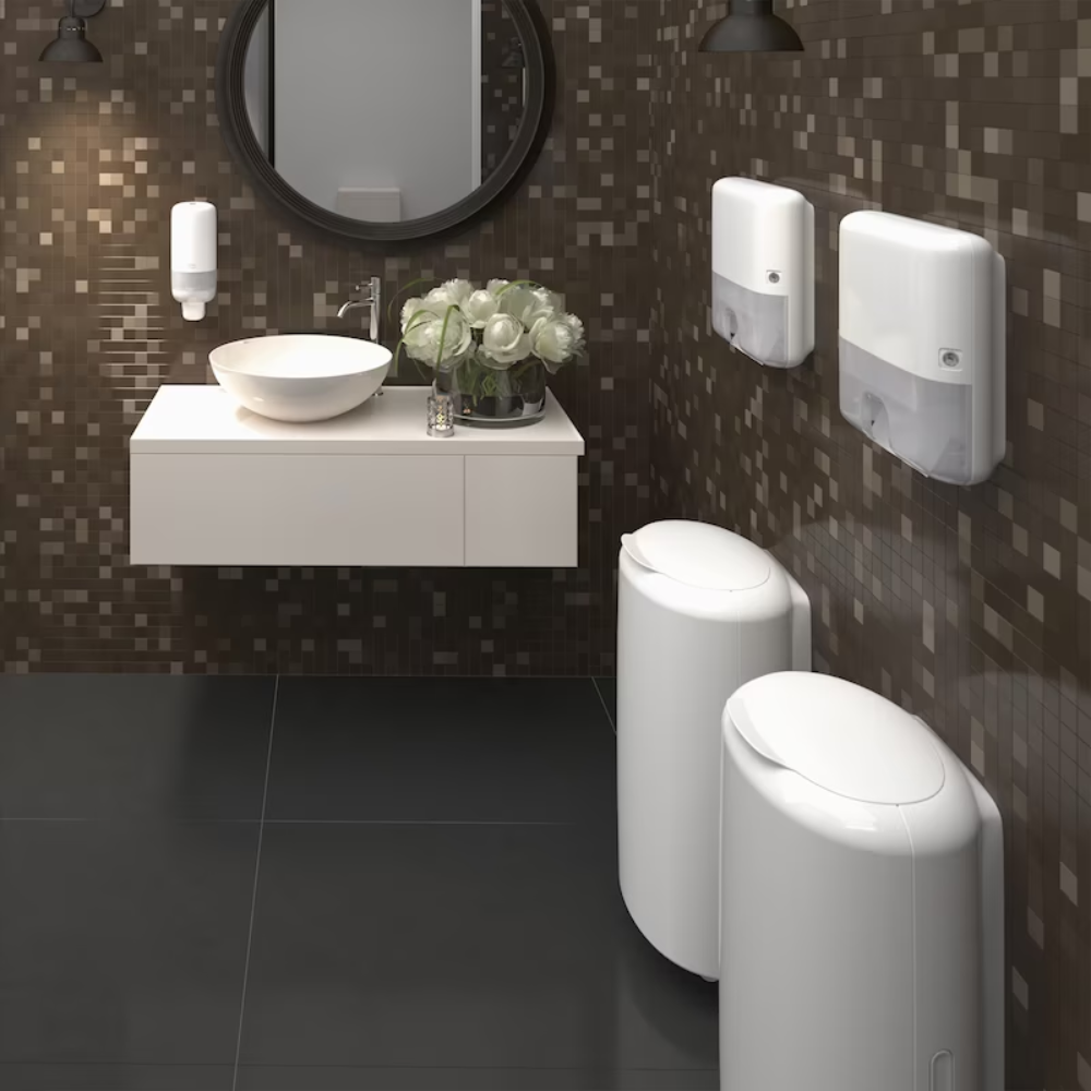 Ein modernes Badezimmer mit Mosaikfliesen an den Wänden, einem runden Spiegel über einem weißen Waschbecken, zwei weißen Handtuchspendern an der Wand mit Tork Xpress® 552100 Mini Spender für Multifold Handtücher Elevation H2 | Packung (1 Stück) von TORK, einem Seifenspender, einer Vase mit weißen Blumen auf der Ablage und zwei weißen Mülleimern auf dem Boden.