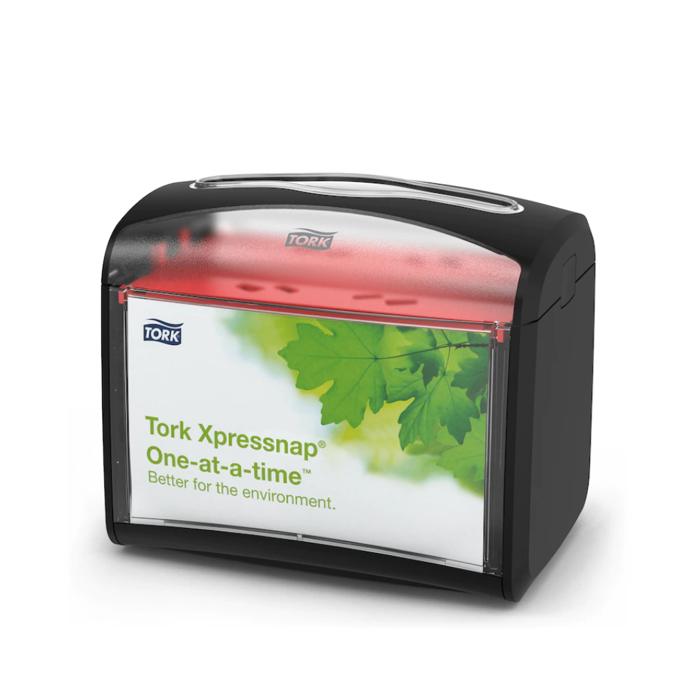 Bild eines Tork Xpressnap® 272611 Tischspenders für Servietten | Karton (4 Packungen) mit schwarzem Gehäuse und transparenter Vorderseite mit TORK-Logo und grünem Blattdesign. Der Text auf der Vorderseite lautet „Tork Xpressnap, eins nach dem anderen, besser für die Umwelt“, ideal, um den Serviettenverbrauch in jeder Umgebung zu reduzieren.