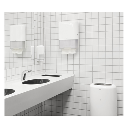 A modern public restroom features a white sink with two faucets, a white tiled wall, and a TORK Tork 100278 extra soft zigzag towels Premium H3 2-ply | Carton (15 packs) dispenser with high-quality 2-ply paper towels. Complemented by a soap dispenser and a trash can, the environment is clean and minimalist.