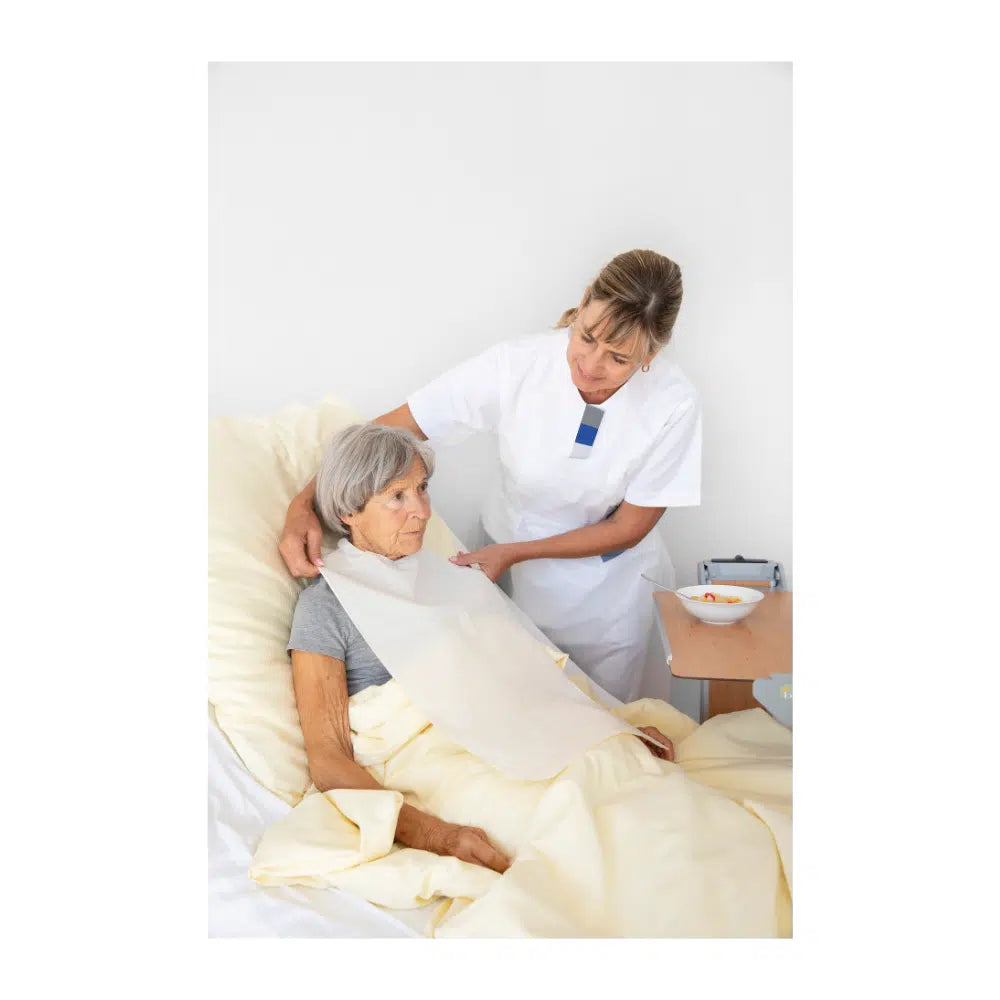 A nurse adjusts the blanket for an elderly woman who is sitting upright in a hospital bed. Both appear attentive and focused. A tray with a bowl and Vala® Fit Tape single-use clothing protection can be seen.