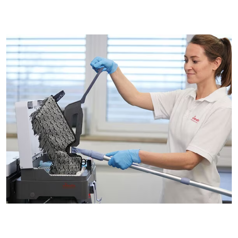 A person with blue gloves and a white shirt is cleaning a textured black surface with the Vileda Professional Swep Duo r-MicroCombi Mop - 50 cm from Vileda GmbH. The scene appears to be indoors, with a large window in the background through which natural light enters the room.