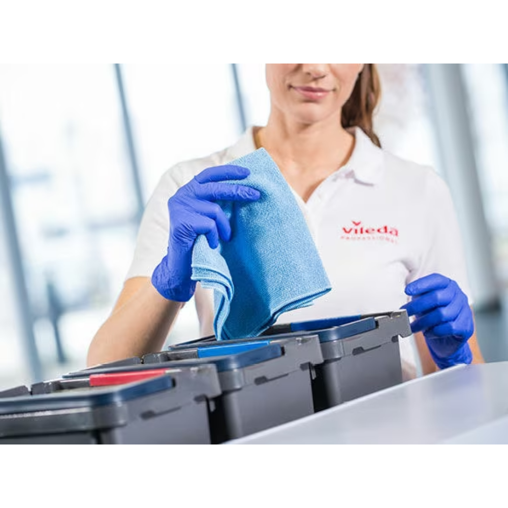 Eine Person mit blauen Handschuhen und weißem Hemd mit Vileda-Aufdruck legt ein Vileda Professional r-MicroTuff Swift Microfasertuch, blau | Packung (5 Tücher) in ein Fach eines Reinigungscaddys mit mehreren Fächern, teilweise mit unterschiedlich farbigen Deckeln.