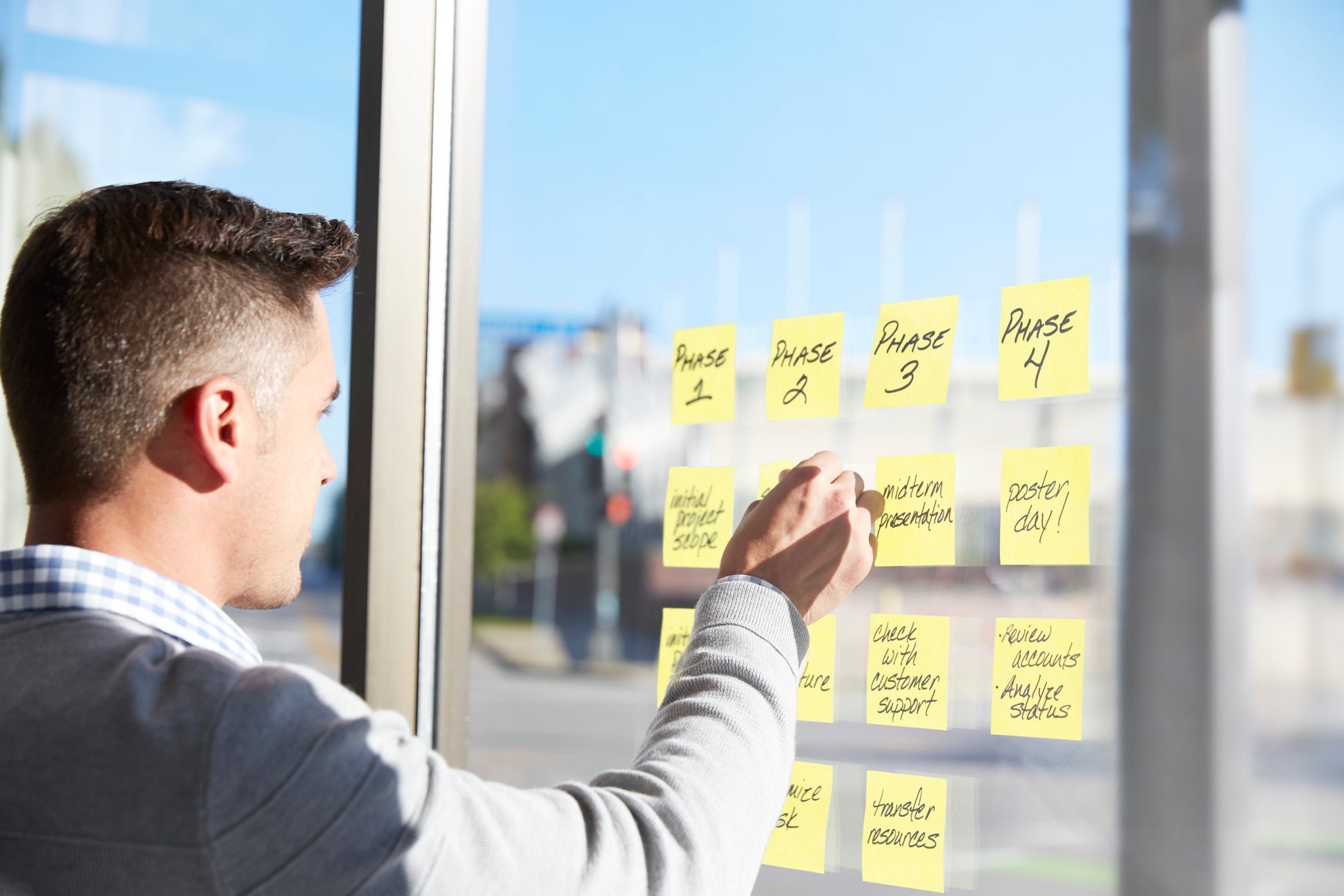 Ein Mann benutzt den 3M Deutschland GmbH Post-it® Z-Notes Dispenser, PRO (Schwarz) und gelbe Post-it® Super Sticky Z-Notes (76x76 mm, 90 Blatt/Block), um Projektaufgaben an einem Glasfenster mit Sonnenlicht und einer Straße draußen zu organisieren.