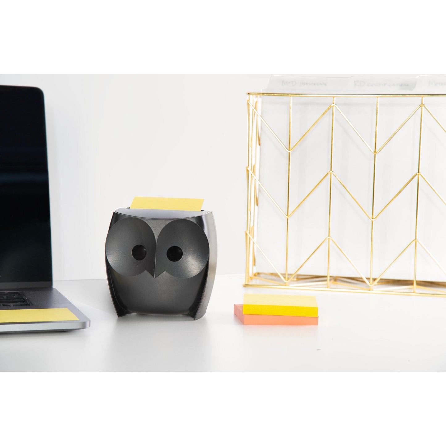 A black, owl-shaped Post-it® Z-Notes Dispenser from 3M Deutschland GmbH stands on a white desk with a pack of Post-it® Super Sticky Z-Notes (45 sheets/block, 2 blocks/pack), next to a laptop and a golden wire organizer.