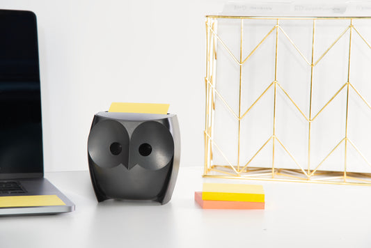 Ein Post-it® Z-Notes Dispenser, Eule, Schwarz von 3M Deutschland GmbH mit gelben Super Sticky Z-Notes (45 Blatt/Block, 2 Blöcke/Packung) steht auf einem weißen Schreibtisch neben einem Aktenordner, zusätzlichen Haftnotizen und einer Laptop-Ecke.