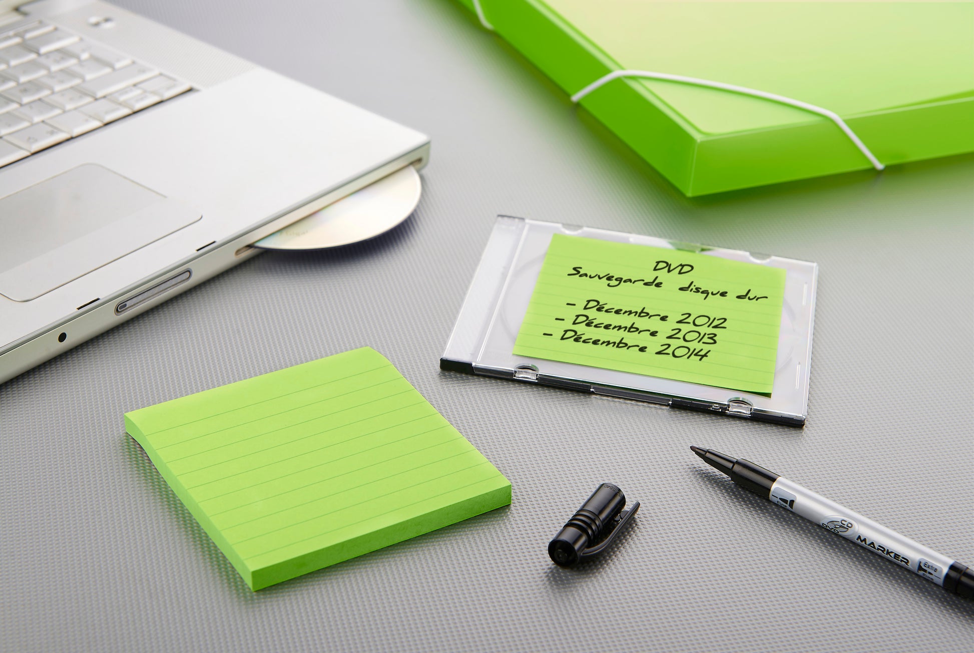 Ein Laptop mit offenem Laufwerk, ein grüner Aktenordner, ein schwarzer Marker ohne Kappe, eine spanisch beschriftete DVD-Hülle und 3M Deutschland GmbH Post-it® Super Sticky Notes im Großformat (101 mm x 101 mm) liegen auf einer grauen Fläche.