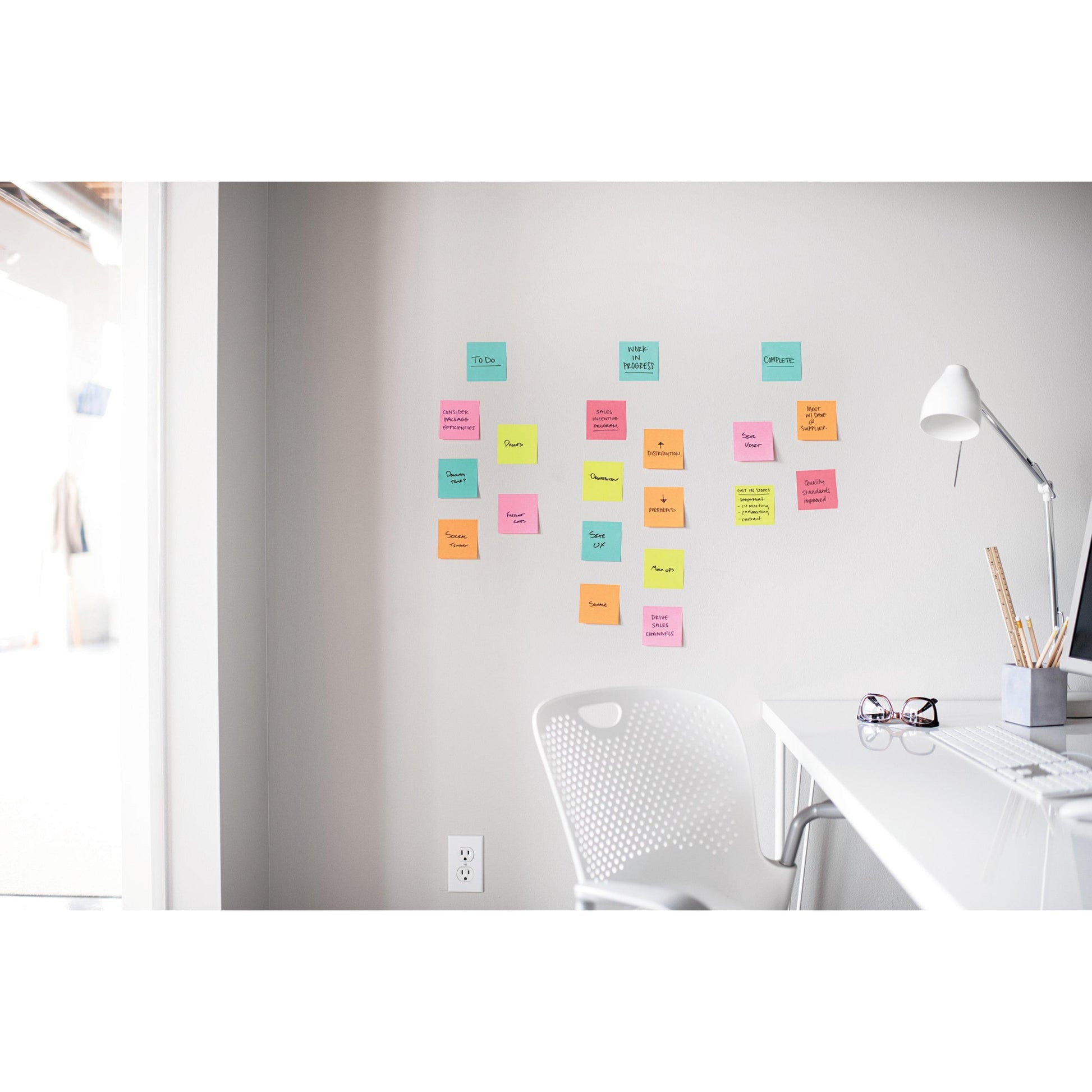 Ein weißer Schreibtisch mit Lampe, Stifthalter und Brille steht neben einem weißen Stuhl. Oben sind in einem hellen Büro Post-it® Super Sticky Notes (47,6 mm x 47,6 mm, 90 Blatt/Block, 100% PEFC) der 3M Deutschland GmbH an der Wand angebracht.