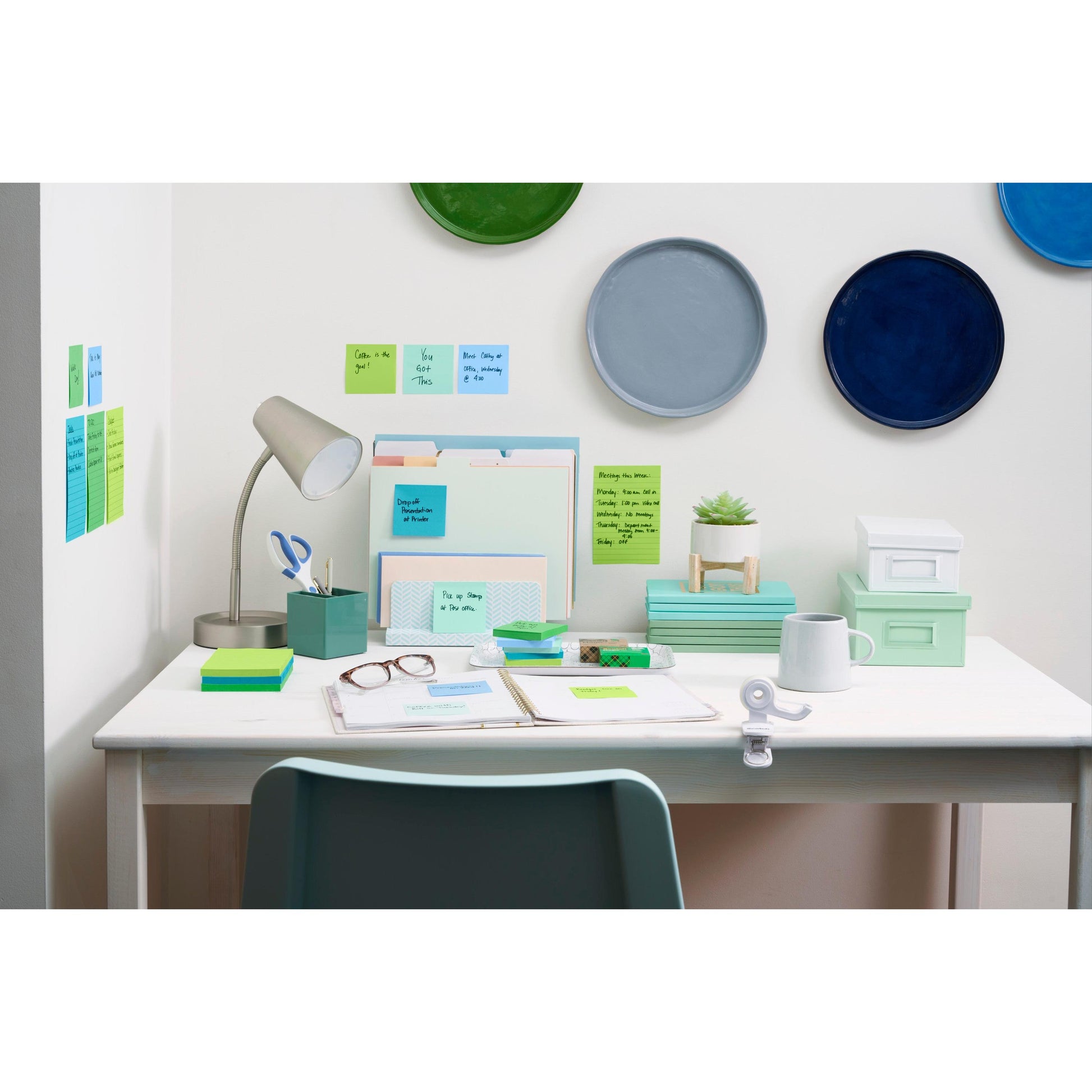Ein aufgeräumter weißer Schreibtisch mit einer Lampe, Ordnern, Notizbüchern, Stiften, Tassen und Gläsern. Die Post-it® Super Sticky Notes (76 mm x 127 mm) der 3M Deutschland GmbH hängen an der Wand mit bunten Tellern. Ein Stuhl steht vor dem Schreibtisch.