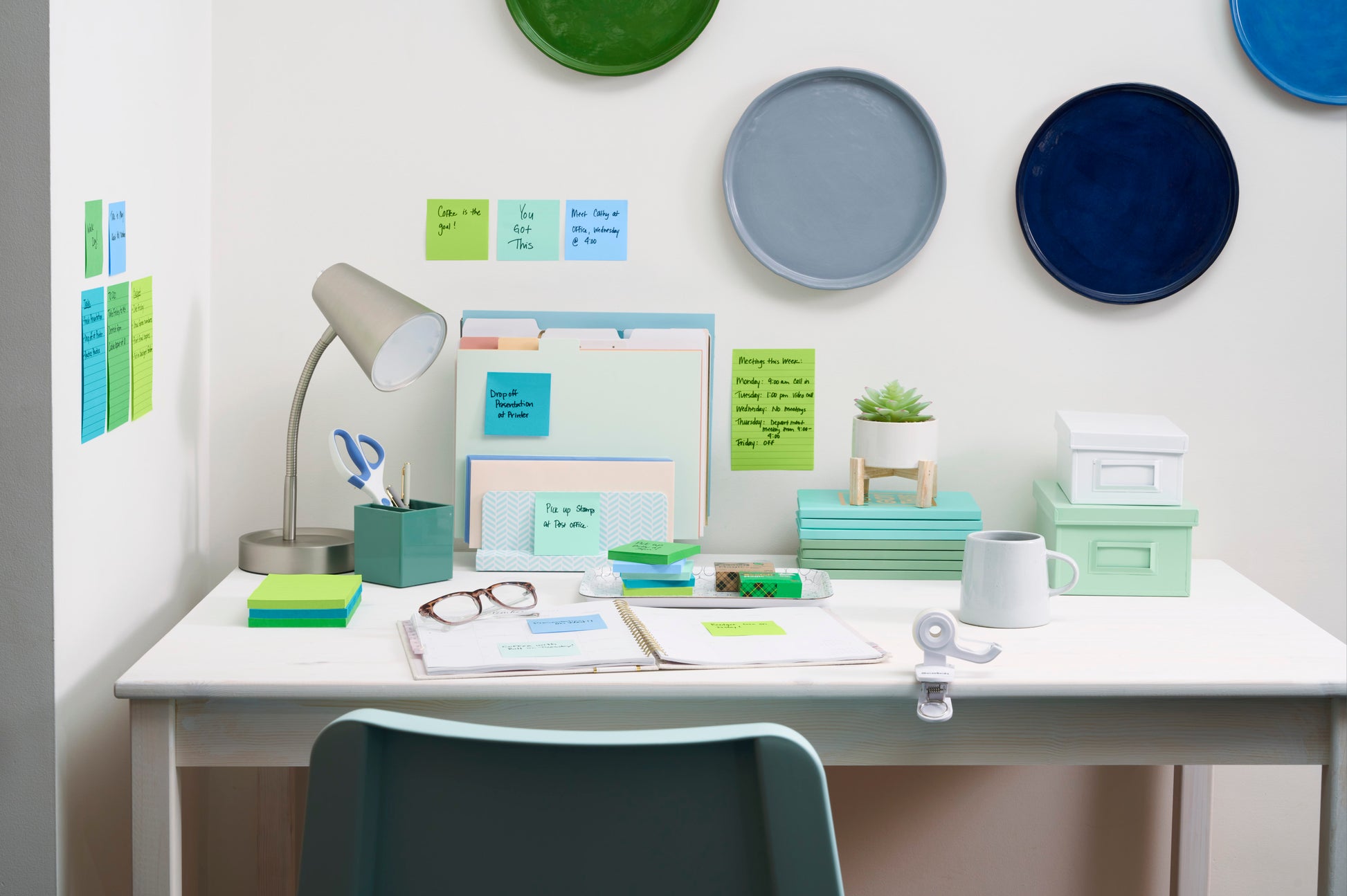 Ein aufgeräumter weißer Schreibtisch mit organisiertem Büromaterial, darunter Post-it® Super Sticky Notes im Großformat (101x101 mm, 70 Blatt/Block, 100% PEFC) von 3M Deutschland GmbH, Ordner, Schreibwaren, eine Lampe, eine Pflanze und Kartons. Buntes rundes Dekor und Haftnotizen an der Wand.