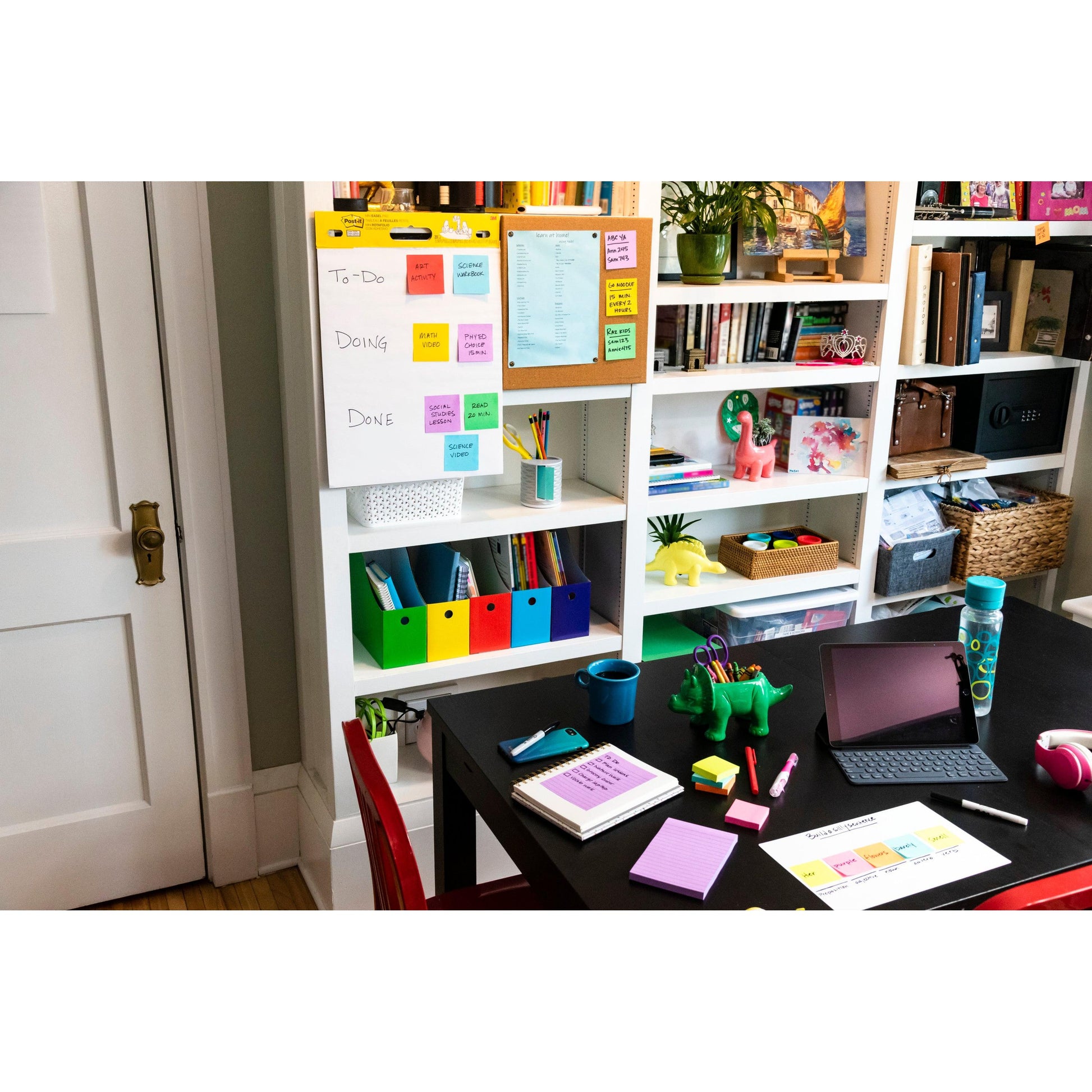 In einem Heimbüro steht ein schwarzer Tisch mit Schreibwaren, einem Tablet und Notizbüchern. Im Hintergrund befinden sich Regale mit Büchern und Ordnern sowie eine Aufgabenliste auf einem Whiteboard mit 3M Post-it® Super Sticky Notes (76x127 mm, 90 Blatt/Block, 100% PEFC).