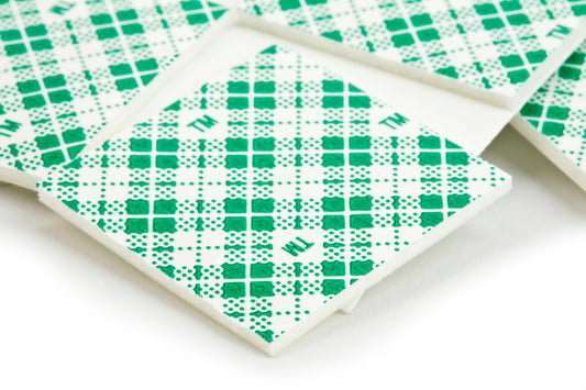 Nahaufnahme von 3M Deutschland GmbH Scotch-FIX™ doppelseitigen Montagequadraten (25 mm x 25 mm, 16 Stück/Packung) in Einzelverpackungen mit grünem Karomuster und verstreuten Buchstaben auf der Verpackung.