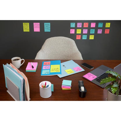 A tidy desk with a laptop, coffee, pens, and office supplies stands in front of a wall covered with rows of 3M Deutschland GmbH Post-it® Super Sticky Notes (47.6mm x 47.6mm, 90 sheets/block, 100% PEFC); an empty chair stands behind the desk.