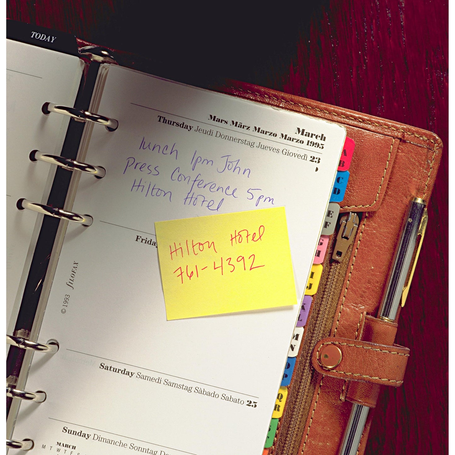 An open leather-bound planner shows handwritten Thursday notes: "Lunch 1 PM John," "Press conference 5 PM Hilton Hotel." On a yellow Post-it® Recycling Notes block from 3M Deutschland GmbH it says: "Hilton Hotel 761-4392." The dividers are visible.