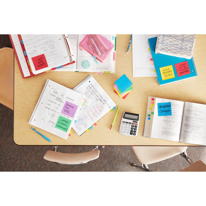 A table in a classroom, neatly arranged with open notebooks, textbooks, a calculator, colored pens, folders, and Post-it® Notes from the Energetic Collection (38mm x 51mm, 100 sheets/block), brand: 3M Deutschland GmbH; three empty chairs surround the table.