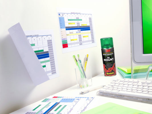 Ein Schreibtisch mit einer Tastatur, Stifte in einer durchsichtigen Halterung, gedruckte Zeitpläne an der Wand und auf dem Schreibtisch, ein Teil eines Monitors und eine Dose 3M™ Scotch® ReMount™ Klebespray (400 ml) der 3M Deutschland GmbH.