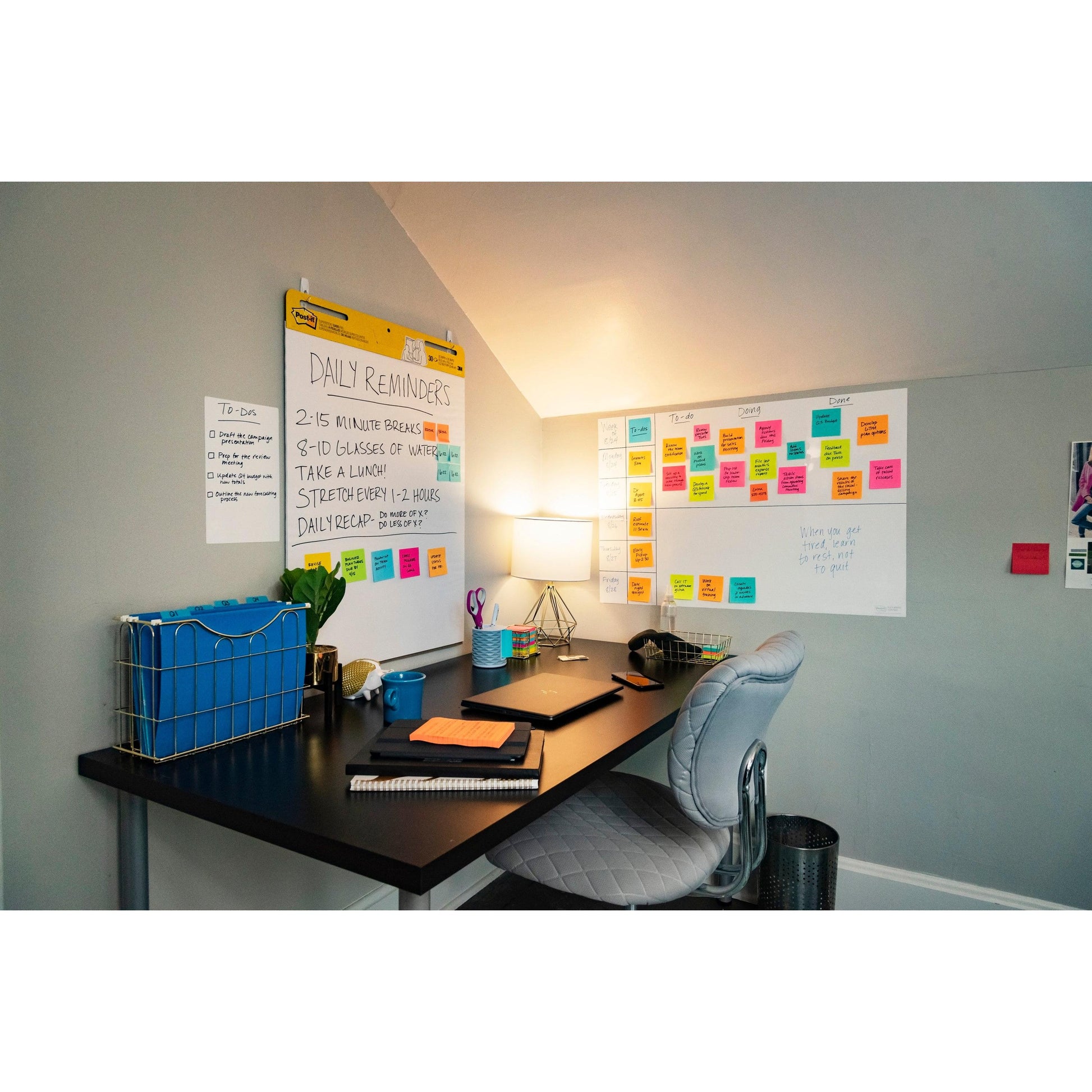 Ein Heimbüro mit schwarzem Schreibtisch, grauem Stuhl, Lampe, Notizbüchern, einem Stifthalter und zwei Post-it® Easy Erase Whiteboard-Oberflächen von 3M Deutschland GmbH, die mit Notizen bedeckt sind. Bunte Haftnotizen und eine To-Do-Liste zieren die Wand.