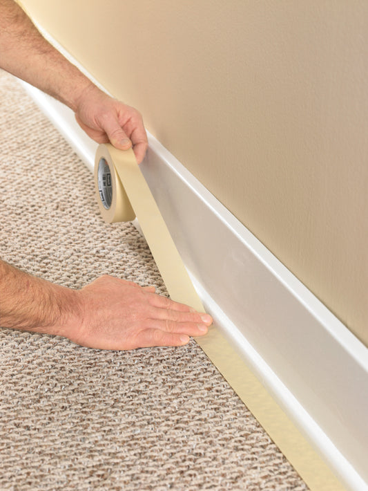 Eine Person verwendet Scotch® Classic Abdeckband (36 mm x 50 m, 1 Rolle) von 3M Deutschland GmbH zum Abkleben der Sockelleiste, wo der Teppich auf die Wand trifft, als Vorbereitung für Maler- oder Renovierungsarbeiten. Enthält 70% PEFC-zertifiziertes Material.