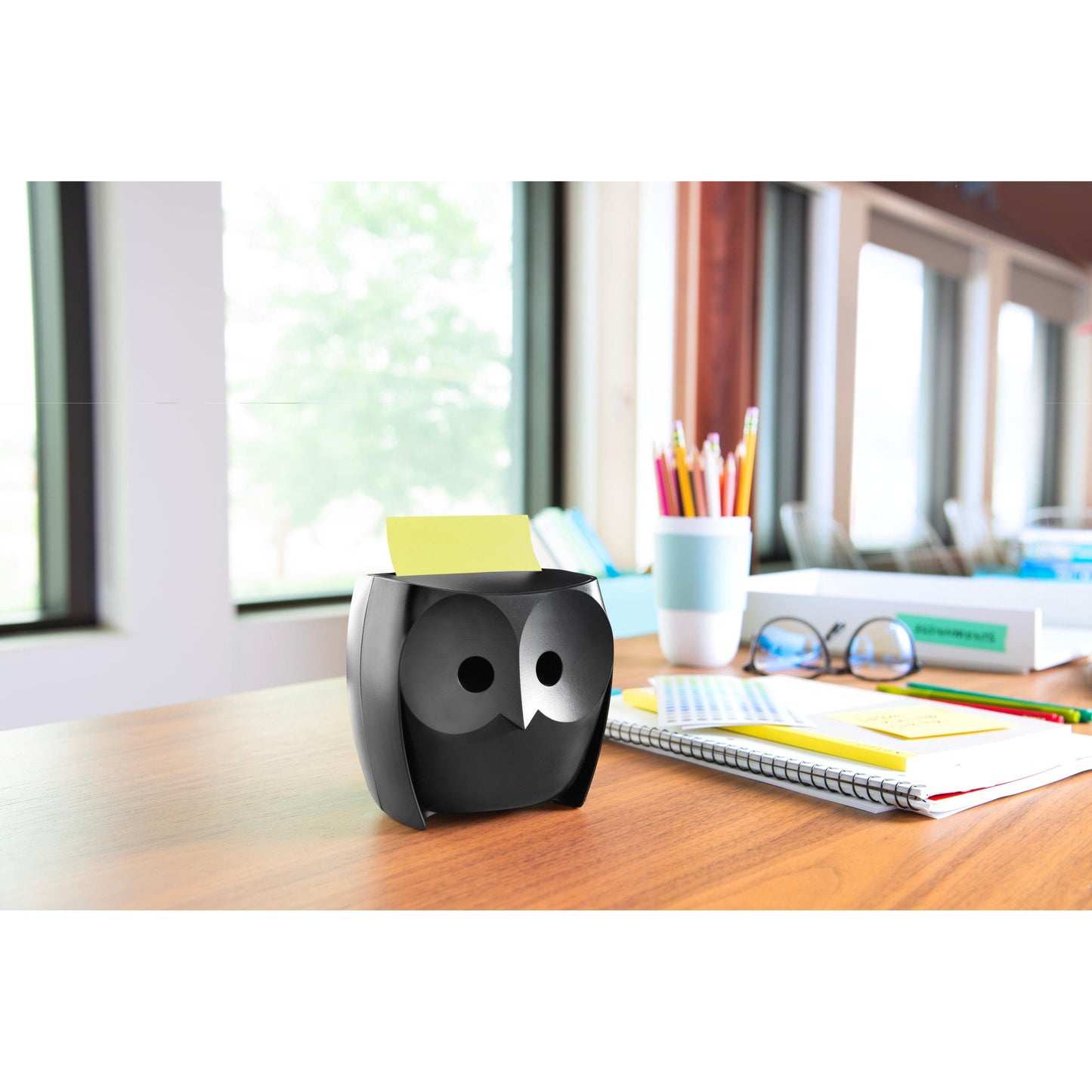 A Post-it® Z-Notes Dispenser, Owl, Black from 3M Deutschland GmbH stands on a wooden table with notebooks, a cup, colored pencils in a cup, and Post-it® Super Sticky Z-Notes (45 sheets/block, 2 blocks/pack); large windows offer a view of trees.
