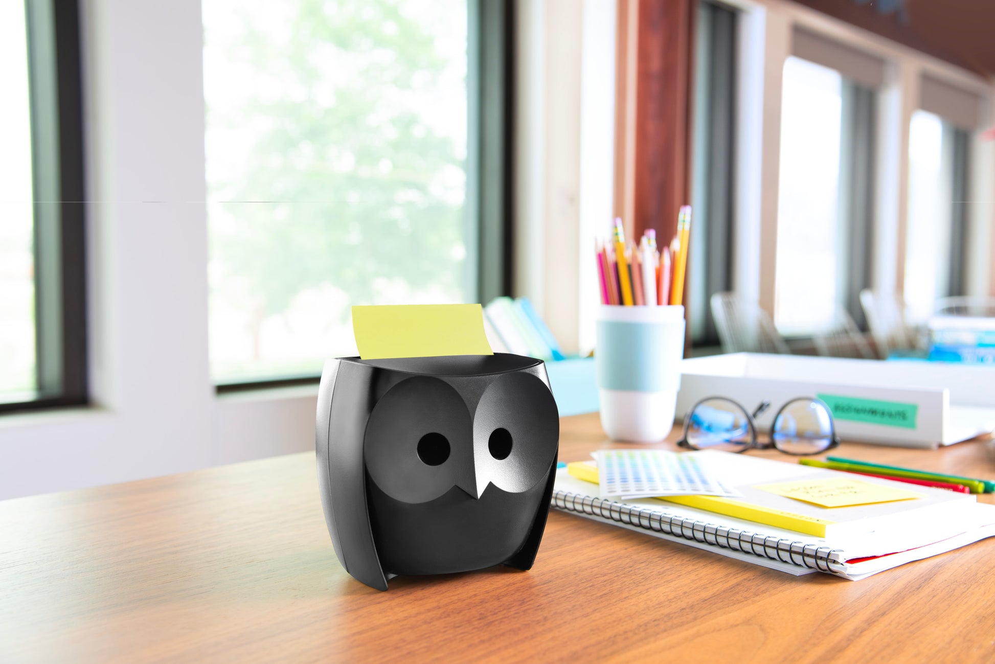 Der 3M Deutschland GmbH Post-it® Z-Notes Dispenser, Eule, Schwarz, mit Post-it® Super Sticky Z-Notes (45 Blatt/Block, 2 Blöcke/Packung), steht auf einem Holztisch neben Notizbüchern und Stiften in einem hell erleuchteten Raum.