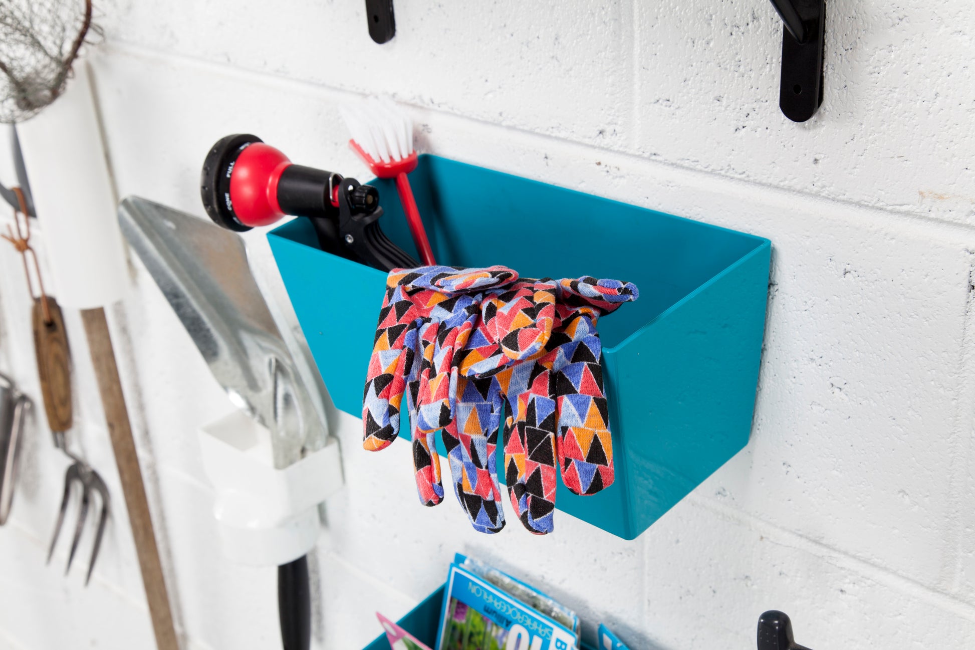 Ein Paar bunte Gartenhandschuhe hängt über einem blauen Eimer, der mit Scotch-FIX™ Extreme doppelseitigem Außenmontageband der 3M Deutschland GmbH an einer weißen Wand befestigt ist. In dem Behälter befinden sich eine Sprühdüse und eine Reinigungsbürste, daneben hängen Gartengeräte.