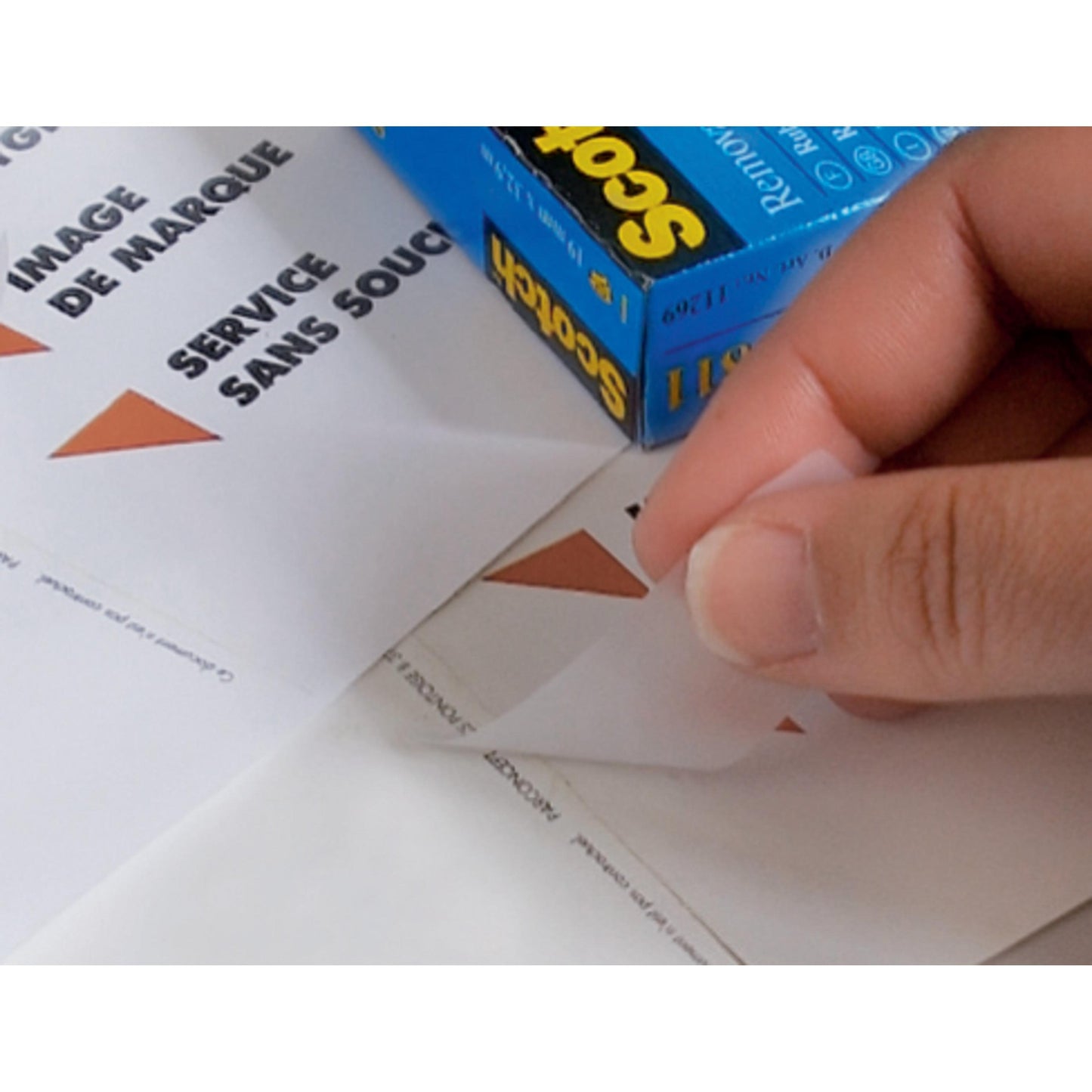 Eine Hand zieht einen dreieckigen Aufkleber von einem Blatt mit der Aufschrift "IMAGE DE MARQUE SERVICE SANS SOUCI" ab, während im Hintergrund eine Schachtel 3M Deutschland GmbH Scotch® unsichtbares Klebeband, ablösbar, 19 mm x 33 m, 1 Rolle | 1 Stück steht.