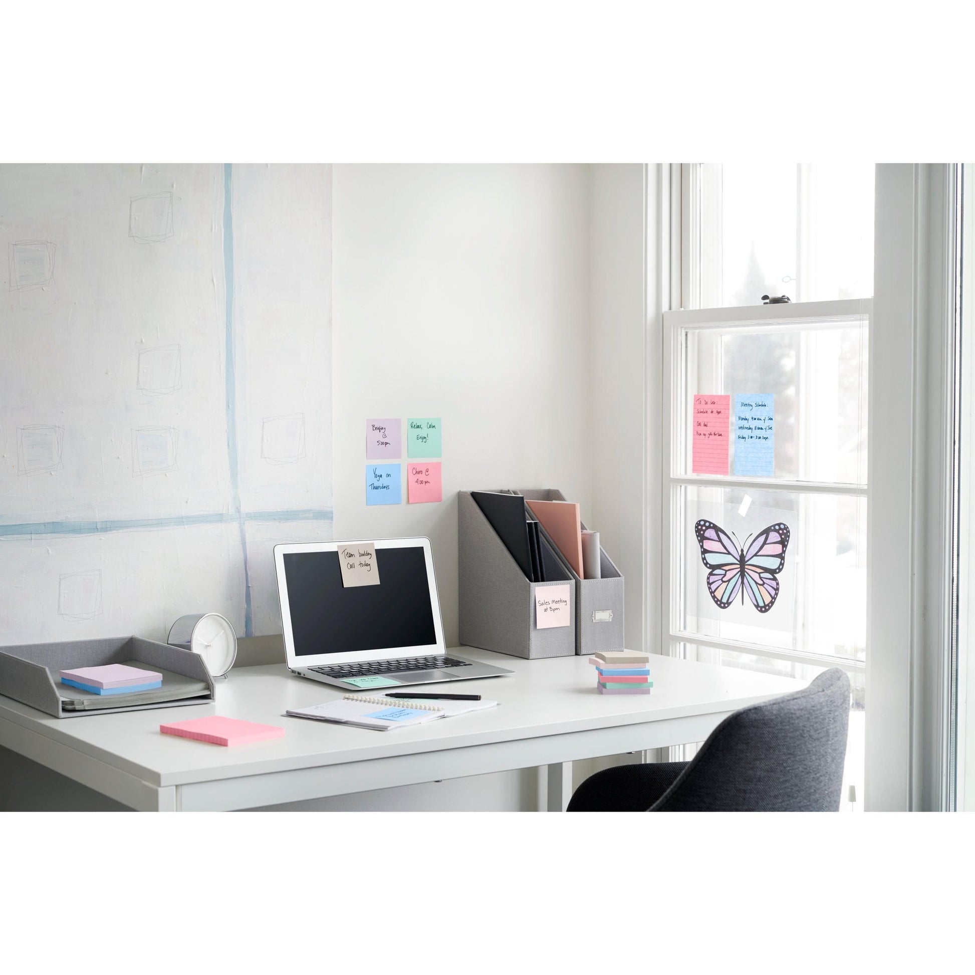 Ein aufgeräumter Bürotisch mit Laptop, Uhr, Notizblock, Aktenordner und 3M Deutschland GmbH Post-it® Super Sticky Notes (47,6x47,6 mm, 90 Blatt/Block, 100% PEFC). Neben dem Schreibtisch befindet sich ein Fenster mit Schmetterlingsdekoration und Haftnotizen in Pastelltönen.