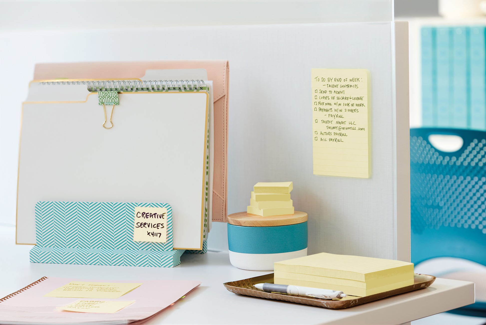 Auf einem aufgeräumten Bürotisch steht der Post-it® Z-Notes Dispenser PRO der 3M Deutschland GmbH in Schwarz mit gelben Post-it® Super Sticky Z-Notes (76x76 mm, 90 Blatt), dazu Aktenordner, ein Stift, ein Behälter mit Holzdeckel und ein blauer Stuhl.