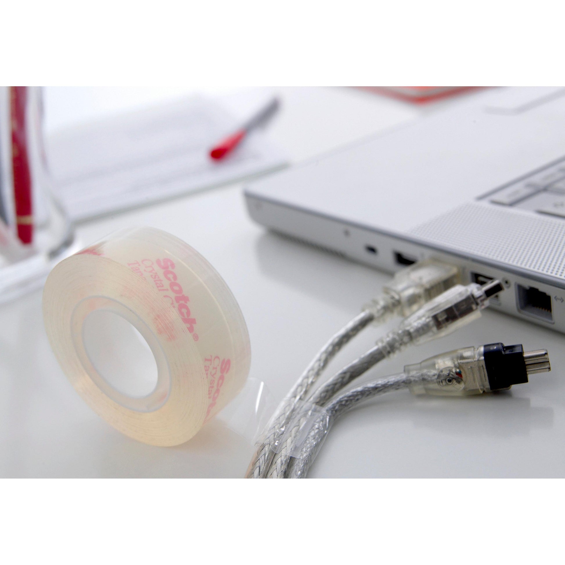 Eine Rolle Scotch® Crystal Klebeband von 3M (19 mm x 33 m, transparent) liegt auf einem weißen Schreibtisch neben zwei durchsichtigen Computerkabeln und einer silbernen Laptop-Ecke, mit einem verschwommenen roten Stift und Papier im Hintergrund.