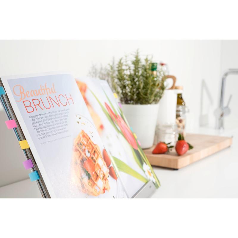An open cookbook with the Post-it® Index Tabs Mini from 3M Deutschland GmbH marks a brunch recipe. In the background, strawberries, a pot with herbs, and kitchen utensils are arranged on a wooden board on a white countertop.