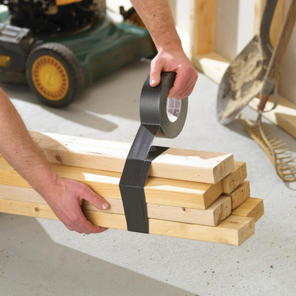 In a workshop, someone secures wooden boards with Scotch® Extremium™ Ultra High Performance Tape DT17 (10 m x 24 mm) by 3M Deutschland GmbH. Nearby are a shovel, a rake, and a lawnmower.